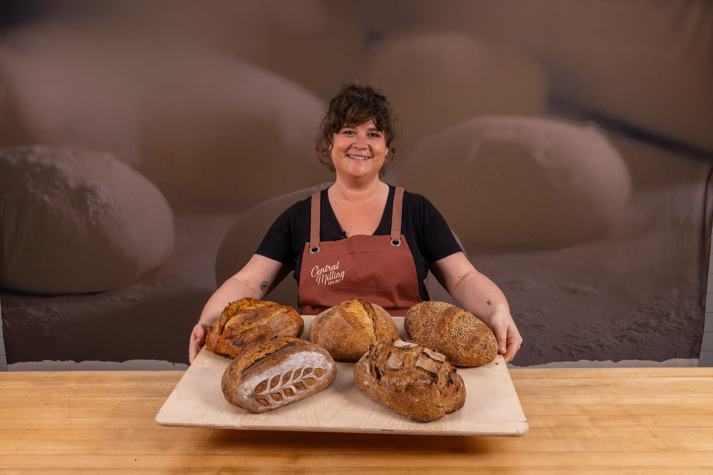Laila smiling in a rust-colored Central Milling apron, holding a wooden board loaded with five decorated sourdough loaves